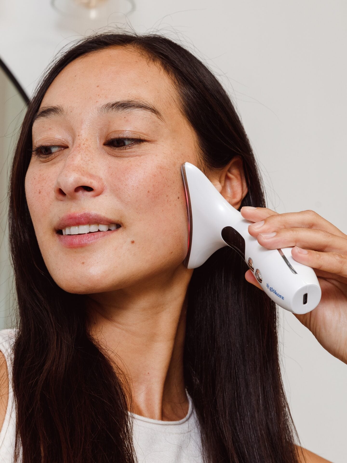 Over-the-shoulder angle of a Black female standing in a bathroom mirror using the Glokore Facial Sculptor on her cheek, soft morning light, countertop skincare products visible, realistic home environment.