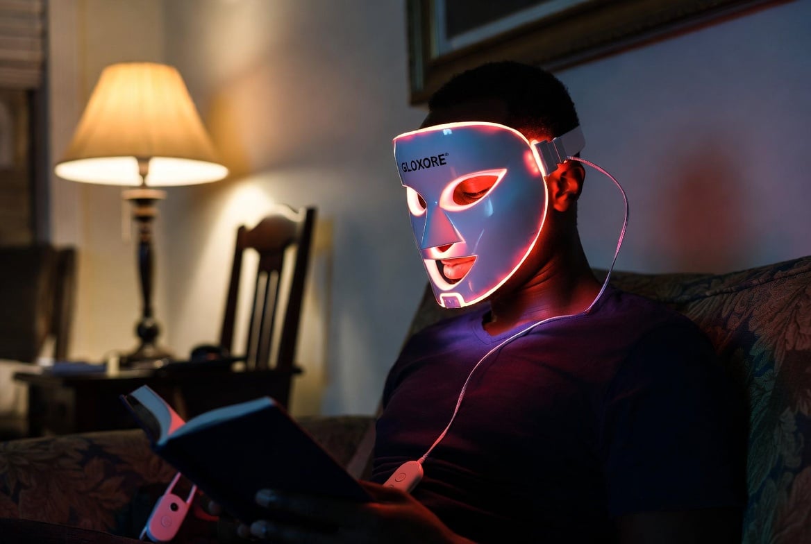 The GLOKORE Wireless LED Light Therapy Mask being used during a candid moment, framed naturally within its environment with slight motion and depth for realism. Black male wearing the glowing mask while reading a book on the couch in a lived-in living room with warm lamp light.