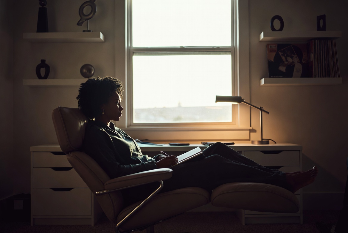 Generational Echoes: A candid, warm-toned photograph in a dining area. A vintage mahogany table is set with a mix of heirloom patterned china and modern dinnerware. Resting near a centerpiece is an old, leather-bound photo album. In the background, on a dark blue-grey accent wall, sits a gallery arrangement of framed family photographs (Black families across generations) and original print art. The lighting is soft and ambient. This shot is intimate, focusing on textures, family history, and personal narrative.