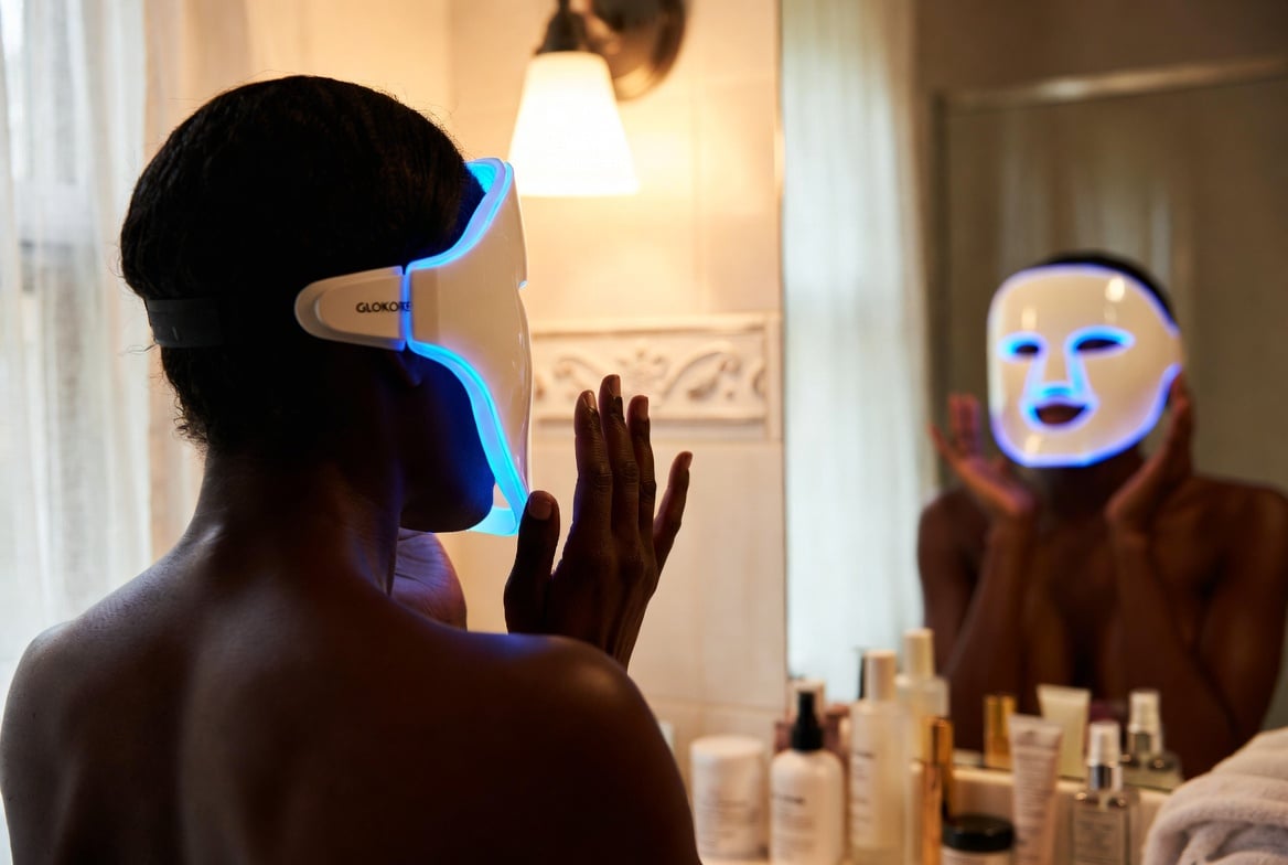 The GLOKORE Wireless LED Light Therapy Mask being used during a candid moment, framed naturally within its environment with slight motion and depth for realism. Black male wearing the glowing mask while reading a book on the couch in a lived-in living room with warm lamp light.